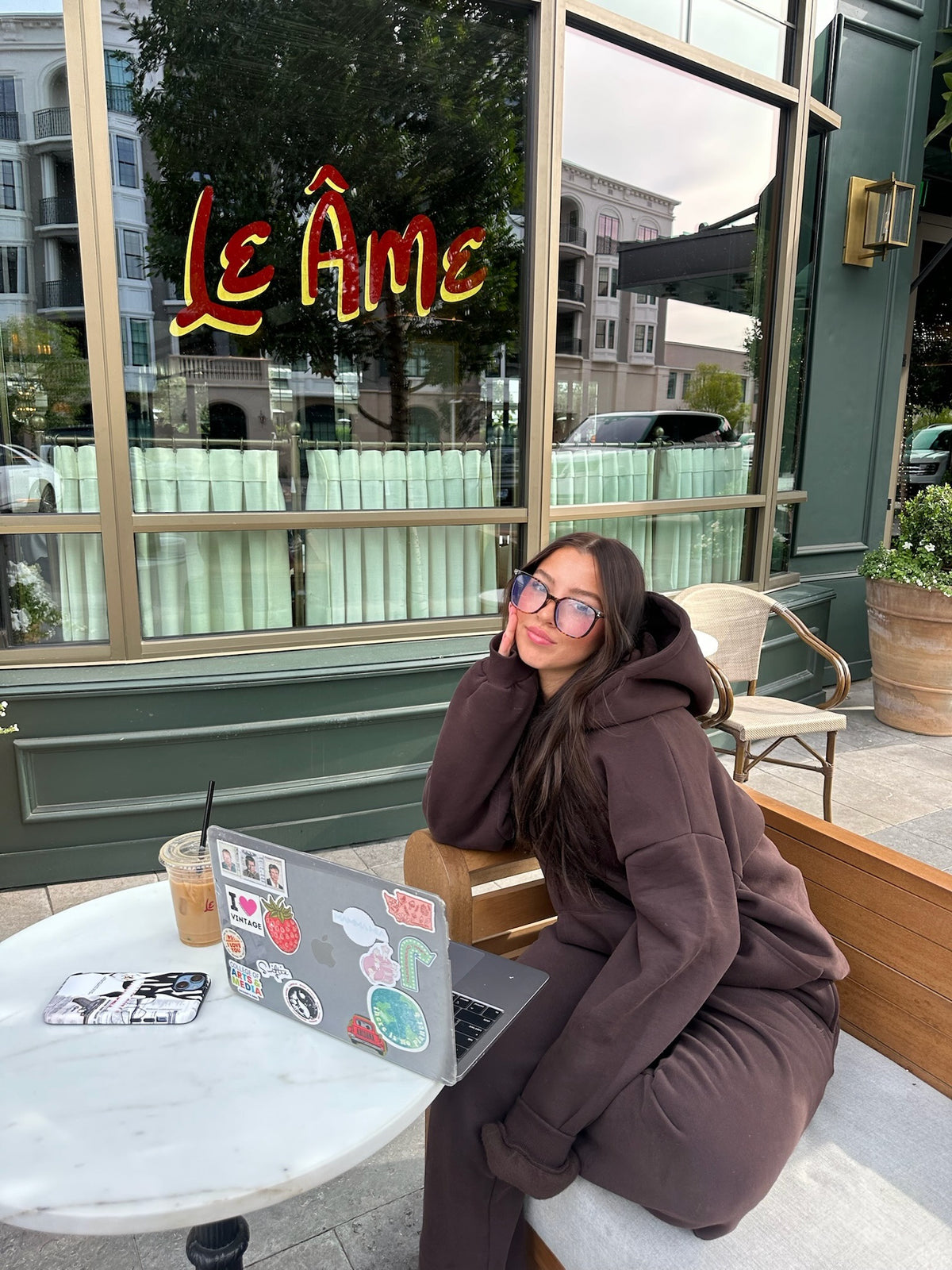 Woman wearing the brown Cascade sweatsuit at a café with a laptop and iced coffee
